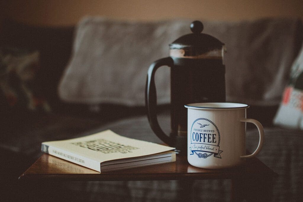 A french press, coffee mug, and book sitting on a coffee table.