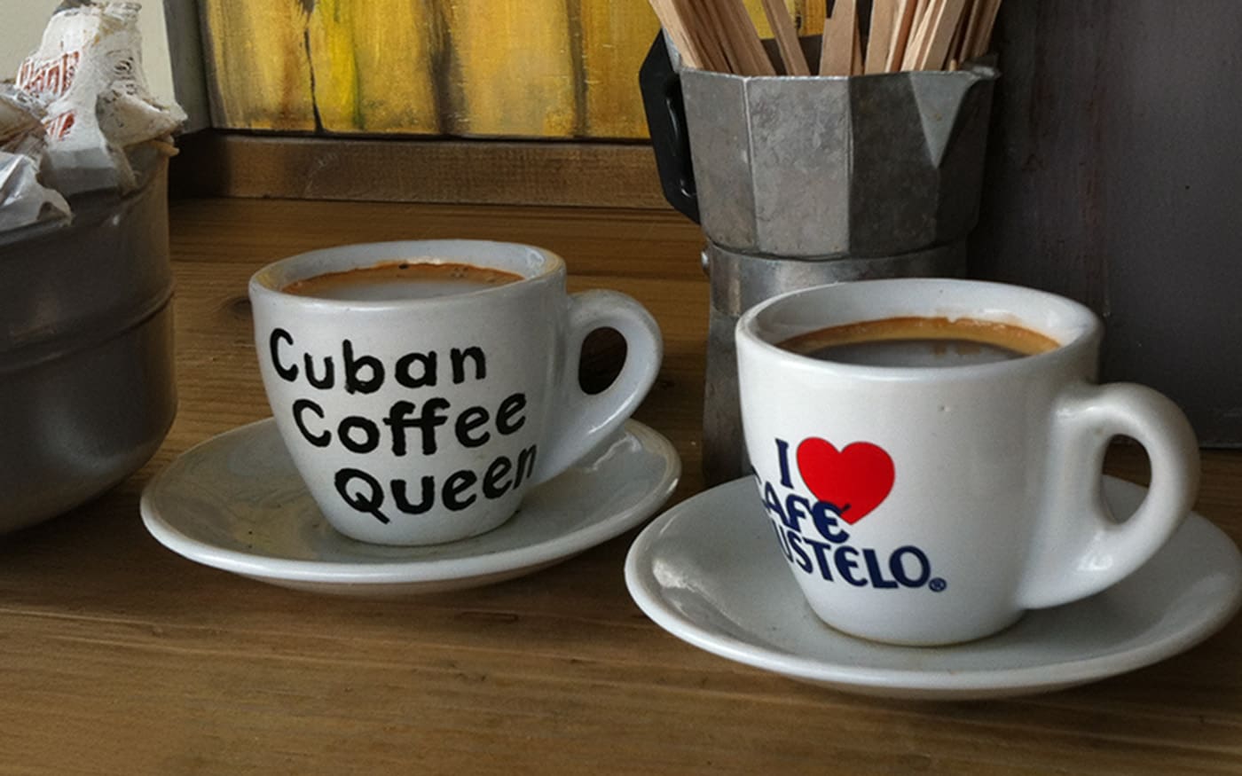 Two white coffee cups on saucers, one labeled 'Cuban Coffee Queen' and the other 'I Love Cafe Bustelo,' filled with Cafecito, set on a wooden table.