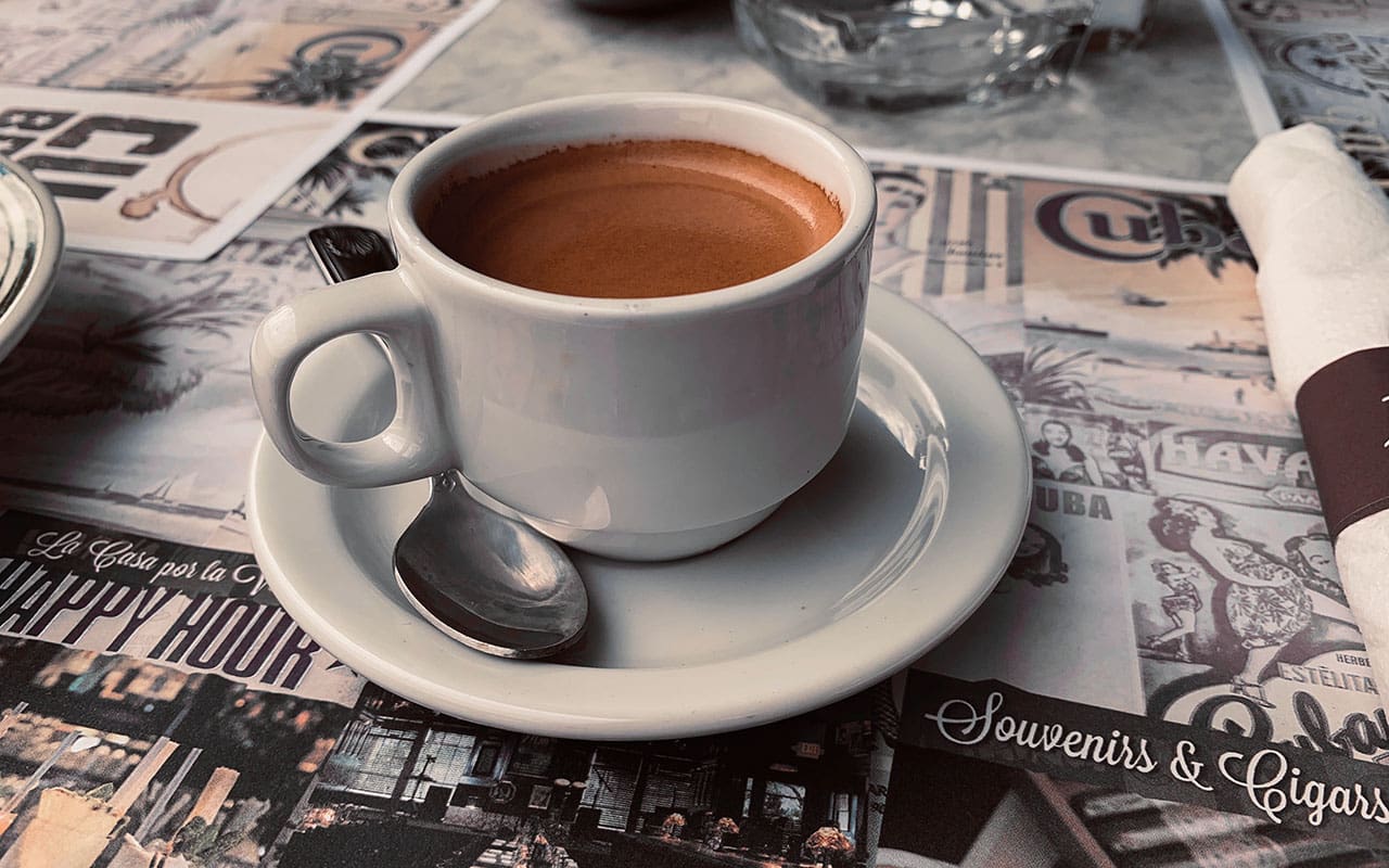 A close-up of a cup of Cafecito served in a white cup with a saucer, set on a table decorated with Cuban-themed paper placemats.