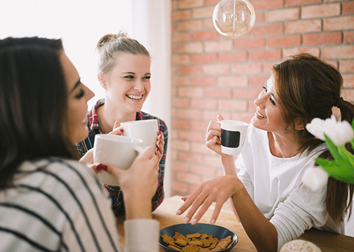 What Is an Americano? Recipe, History, and How It Differs From Drip Coffee Three people sharing coffee around a wooden table with cookies and tulips, set in a cozy indoor space.
