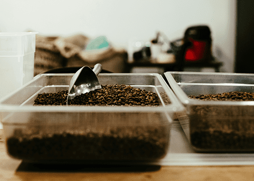 Two Clear Plastic Containers Filled With Roasted Coffee Beans, One With A Metal Scoop Resting On Top.