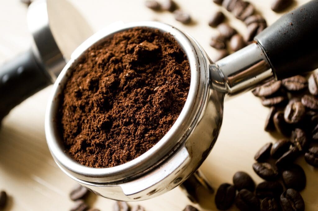 Portafilter filled with freshly ground coffee, surrounded by whole beans on a light countertop.