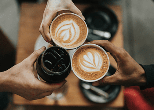 Three hands raising drinks in a toast—two lattes with leaf-shaped latte art and one iced beverage in a glass, over a café table with plates and silverware.