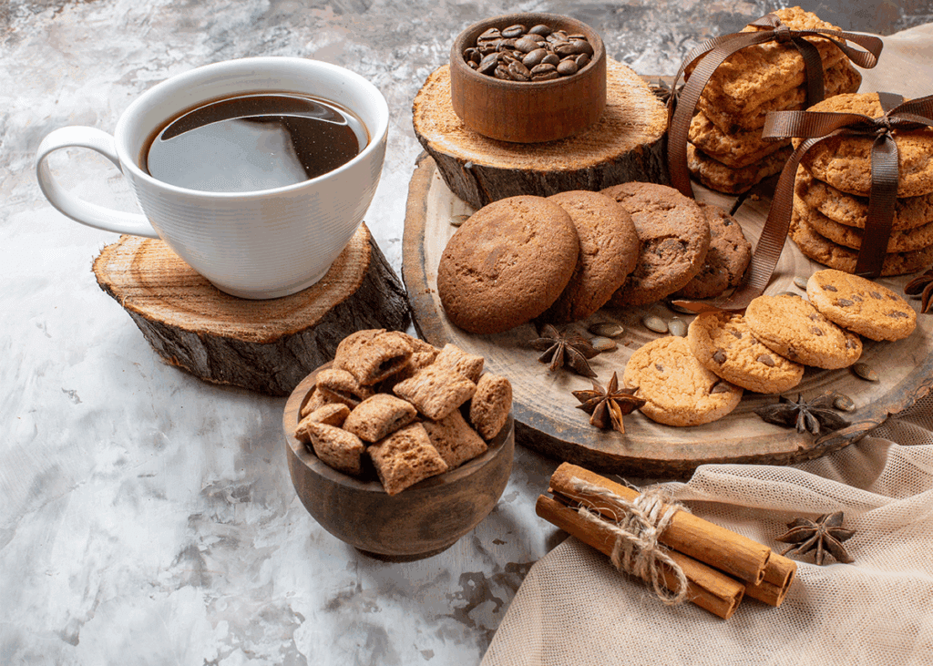 Rustic coffee setup with a white ceramic cup on a wood-slice coaster, surrounded by assorted cookies, coffee beans, and cozy spices on a textured surface