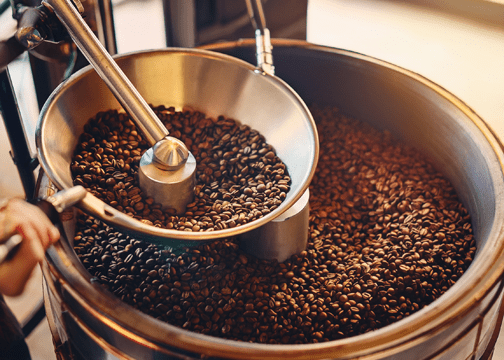 Advanced Coffee Fermentation Techniques Most Roasters Still Don't Know About Large metal coffee roasting drum filled with roasted coffee beans, featuring a mechanical arm with a scoop used for stirring or transferring.