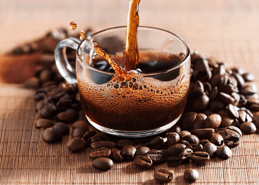 Coffee being poured into a clear glass cup, surrounded by roasted beans on a bamboo mat, with bubbles forming on the surface.