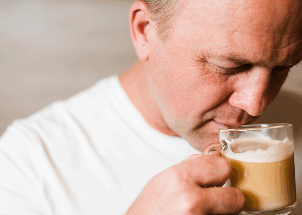 Close-up of a person in a white shirt holding a glass mug of frothy coffee near his face, eyes closed as he inhales the aroma. 