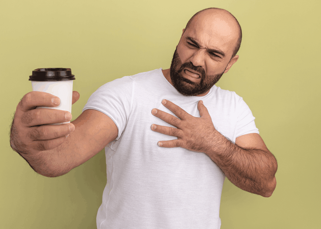A man in a white t-shirt holds a disposable coffee cup with a black lid, grimacing as he places one hand on his chest. His expression conveys discomfort and concern, as if he's questioning how does coffee affects heart health.