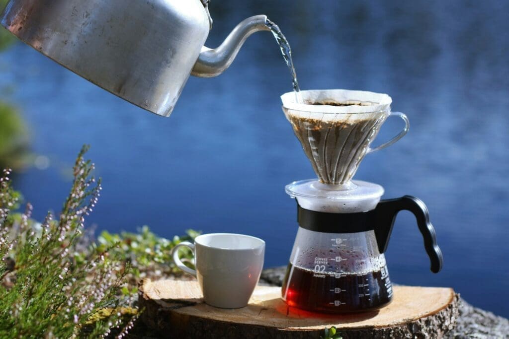 Outdoor pour-over coffee setup with hot water being poured into a glass dripper over a carafe, beside a ceramic cup on a wooden surface near a lake.