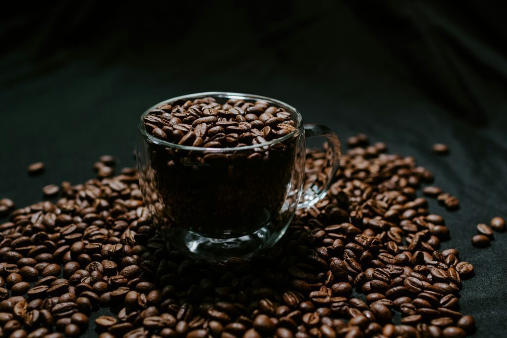 A Clear Glass Cup Filled With Roasted Coffee Beans Sits On A Surface Covered In More Beans, With Dramatic Lighting Highlighting Their Rich Brown Color And Glossy Texture.