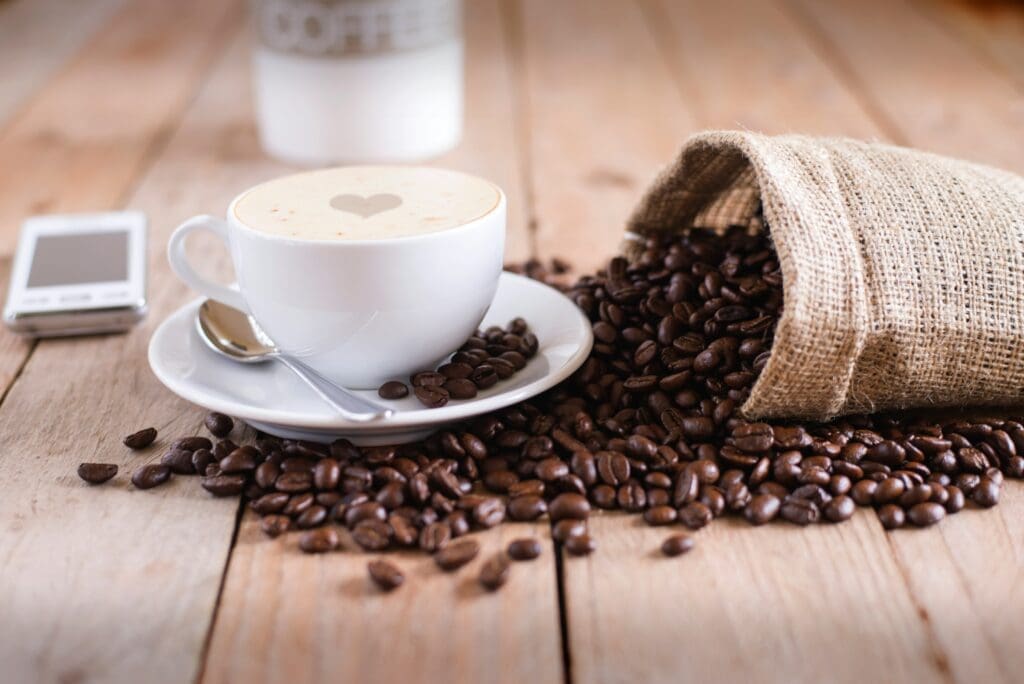 Coffee cup with heart foam on saucer, surrounded by beans spilling from a burlap sack.