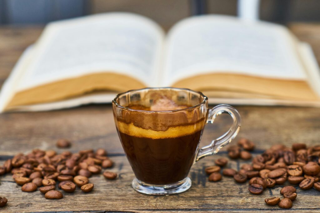 Glass Cup Of Espresso On Rustic Wood With Scattered Coffee Beans, An Open Book In The Background.