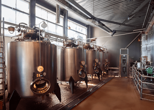 Interior of an industrial facility with stainless steel fermentation tanks, overhead piping, and a control panel, used for large-scale coffee fermentation.